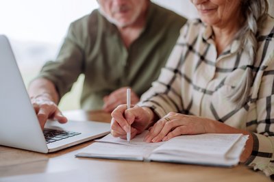 Älteres Paar arbeitet am Laptop, Frau schreibt in Notizbuch, Mann zeigt auf den Bildschirm.