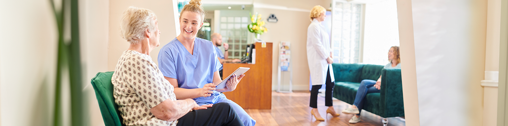 Pflegerin spricht mit älterer Frau auf grünem Sofa in heller Klinik, andere Personen im Hintergrund.