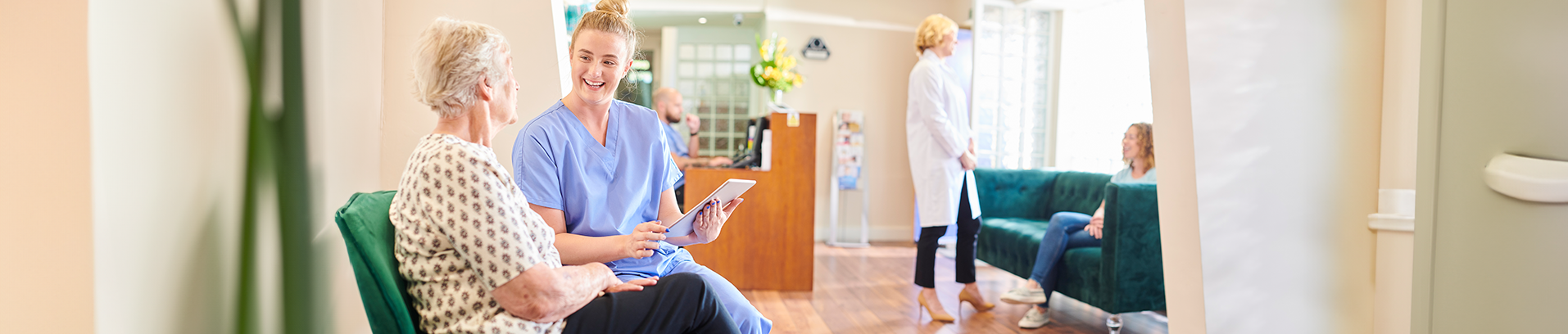 Pflegerin spricht mit älterer Frau auf grünem Sofa in heller Klinik, andere Personen im Hintergrund.