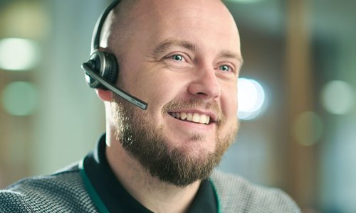 Lächelnder Mann mit Glatze und Bart trägt ein Headset im modernen Büro, unscharfer Hintergrund.