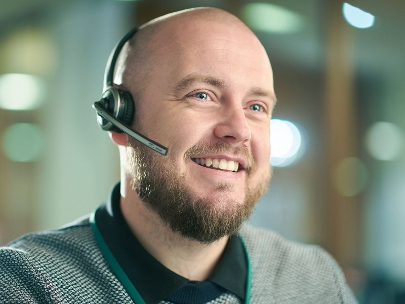 Lächelnder Mann mit Glatze und Bart trägt ein Headset im modernen Büro, unscharfer Hintergrund.