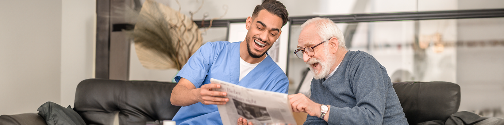 Pflegekraft und älterer Mann lachen und lesen Zeitung auf dunklem Sofa, heller Raum im Hintergrund.