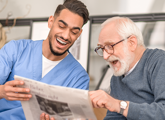 Pflegekraft und älterer Mann lachen und lesen Zeitung auf dunklem Sofa, heller Raum im Hintergrund.