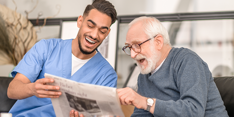 Pflegekraft und älterer Mann lachen und lesen Zeitung auf dunklem Sofa, heller Raum im Hintergrund.