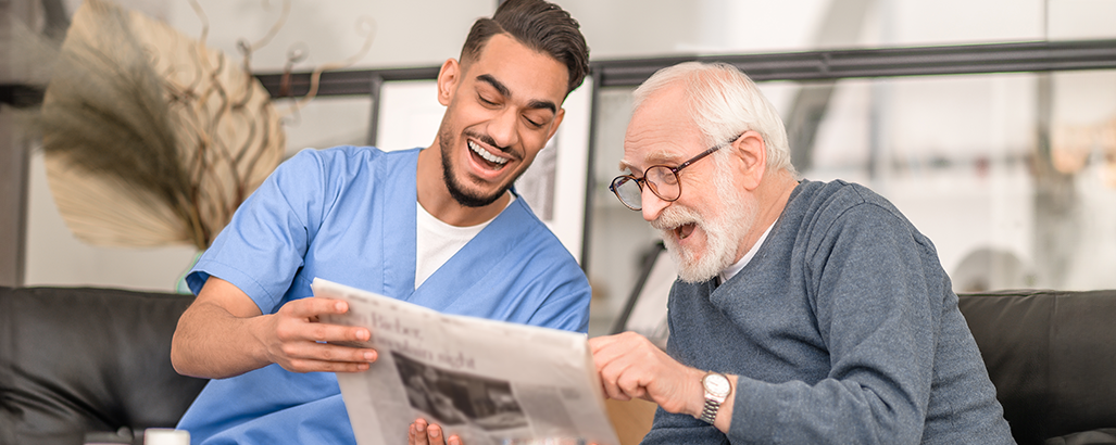 Pflegekraft und älterer Mann lachen und lesen Zeitung auf dunklem Sofa, heller Raum im Hintergrund.