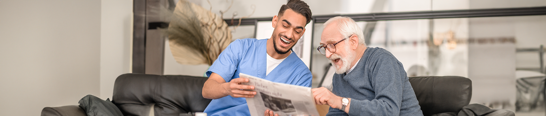 Pflegekraft und älterer Mann lachen und lesen Zeitung auf dunklem Sofa, heller Raum im Hintergrund.